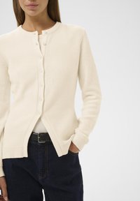 Cardigan lavorato a maglia color crema con scollo rotondo, chiusura frontale con bottoni, polsini a coste e dettaglio di orlo aperto, abbinato a jeans scuri.