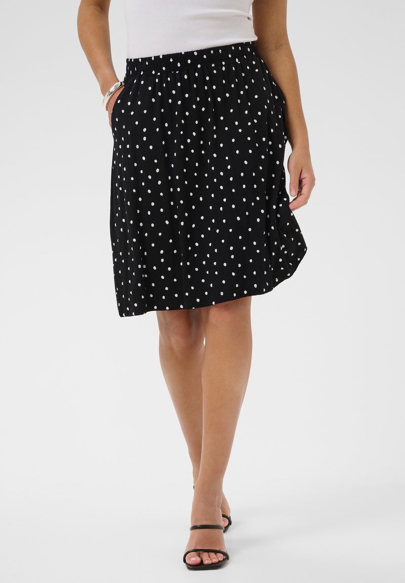 Femme portant une jupe noire à pois blancs, un haut blanc sans manches et des talons noirs ouverts à l'avant, debout avec une main dans la poche.