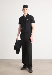 Czarna koszulka polo z akcentami w kolorze turkusowym, połączona z czarnymi spodniami i butami. Model trzyma czarną kurtkę; minimalistyczny design.