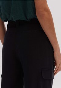 Un pantalon noir présente une coupe décontractée avec des poches latérales et une texture lisse. La ceinture est équipée d'un passant pour une meilleure ajustabilité.