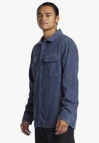 Camisa de pana azul marino con cierre de botones en la parte delantera, dos bolsillos en el pecho, cuello y mangas largas. Presenta un acolchado vertical y un ajuste relajado.
