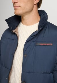 Uomo che indossa un piumino blu navy sopra un maglione bianco a treccia, con particolare attenzione al colletto della giacca e alla patch del logo.