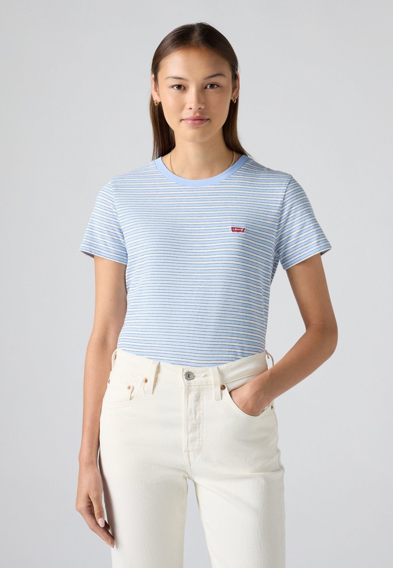 T-shirt a righe blu e bianche realizzato in tessuto morbido, con scollatura rotonda e una piccola toppa con logo rosso sul petto. Abbinato a jeans bianchi.