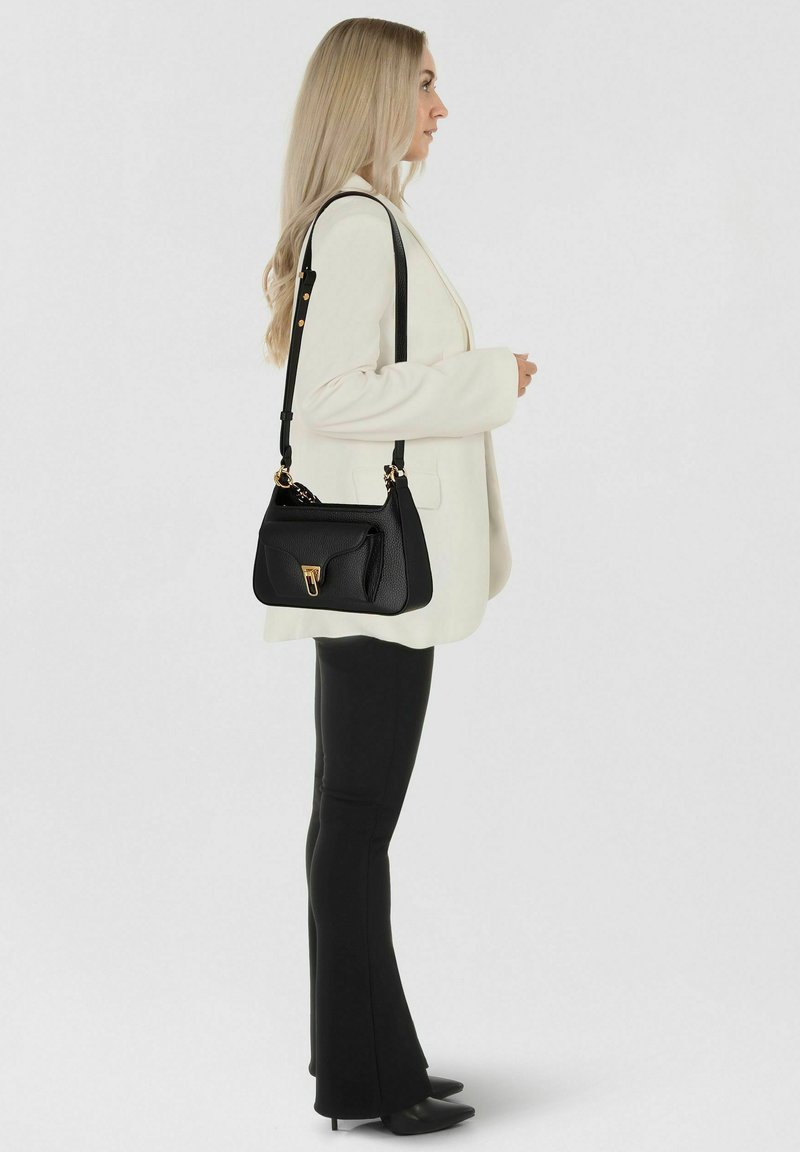 Bolso de cuero negro con forma estructurada, detalles de herrajes dorados, un bolsillo frontal y una correa desmontable. Llevado con un blazer crema.