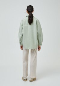 Camicia oversized verde chiaro con bottoni e maniche lunghe, in tessuto morbido; abbinata a pantaloni beige a gamba larga e scarpe beige senza lacci.