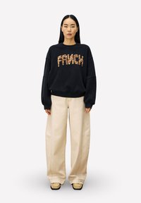 Zwarte sweatshirt met "FRANCH" in gestructureerde bronzen letters, gecombineerd met crèmekleurige wijde broek en tan schoenen, wat zorgt voor een ontspannen look.
