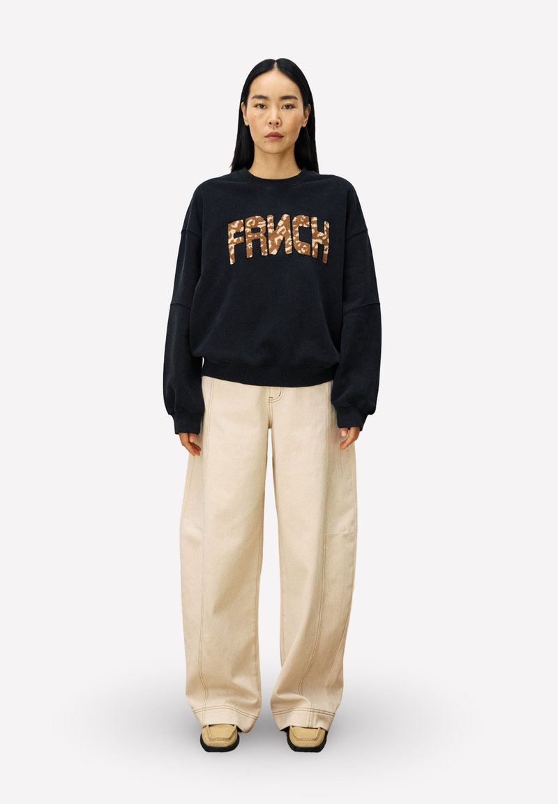 Zwarte sweatshirt met "FRANCH" in gestructureerde bronzen letters, gecombineerd met crèmekleurige wijde broek en tan schoenen, wat zorgt voor een ontspannen look.