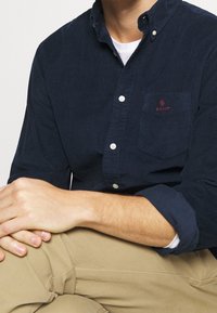 Camicia button-up in corduroy blu navy con taschino e logo rosso. Le mani del modello poggiano su pantaloni khaki, mettendo in risalto la trama e la vestibilità della camicia.