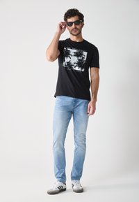 T-shirt en coton noir avec un graphique de palmier, jean bleu clair et baskets grises avec des rayures noires et blanches, portées avec des lunettes de soleil.