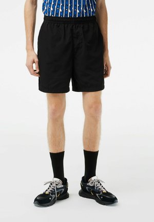Shorts noirs, arrivant jusqu'aux genoux, avec une taille élastique et une texture lisse. Portés avec des chaussettes noires et des baskets au design multicolore.