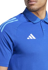 Blaues Polohemd mit Kragen, ausgestattet mit dem weißen Adidas-Logo und drei hellblauen Streifen auf der Schulter. Strukturstoff. Mit Knopfkraagen.