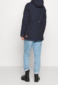 Osoba ubrana w granatową kurtkę z kapturem, jasnoniebieskie jeansy podwinięte na nogawkach oraz czarne buty z sznurówkami, stojąca na tle czystego białego tła.