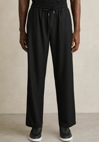 Pantaloni negri, cu dungi, cu talie elastică și �șnur. Cu o croială ușor largă, prevăzuți cu buzunare laterale și asortați cu adidași negri.