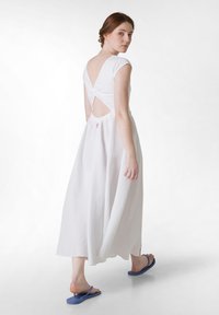 Rochie maxi albă cu decupaj rotund la spate și detaliu încâlcit, cu o croială lejeră și talie elastică. Asortată cu sandale flip-flop albastre.