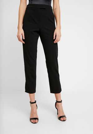 Woman wearing slim-fit black cropped trousers paired with black high-heeled ankle-strap sandals, standing on a plain white background.