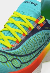 Φωτεινό τυρκουάζ αθλητικό παπούτσι Saucony με κορδόνια νέον κίτρινου χρώματος, πορτοκαλί λεπτομέρειες και κοντινή όψη με υφή και σχέδιο στο πάνω μέρος.