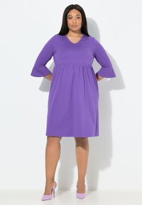 Fioletowa sukienka do kolan z dekoltem w kształcie litery V, rękawami do łokcia, marszczonym pasem i gładką fakturą. Noszona z pasującymi jasnofioletowymi szpilkami.