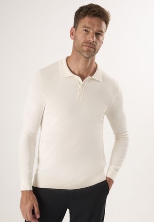Homme aux cheveux courts marron portant un polo blanc à manches longues et un pantalon noir sur un fond uni clair.