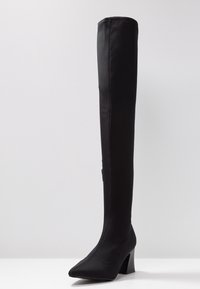 Botte noire mi-cuisse en tissu extensible, dotée d'un bout pointu et d'un talon haut géométrique. Texture lisse avec des détails de couture minimaux.