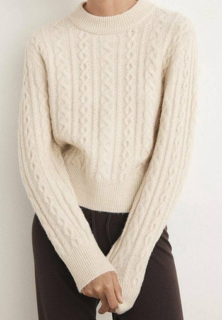 Personne portant un pull en maille torsadée couleur crème à manches longues, associé à un pantalon foncé, debout les mains jointes devant.