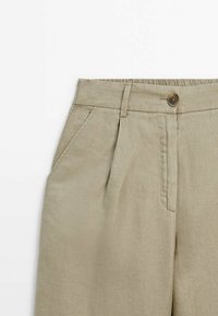 Zone de taille beige clair d’un pantalon avec fermeture par bouton, passants pour ceinture, pli avant et poche latérale sur fond blanc uni.