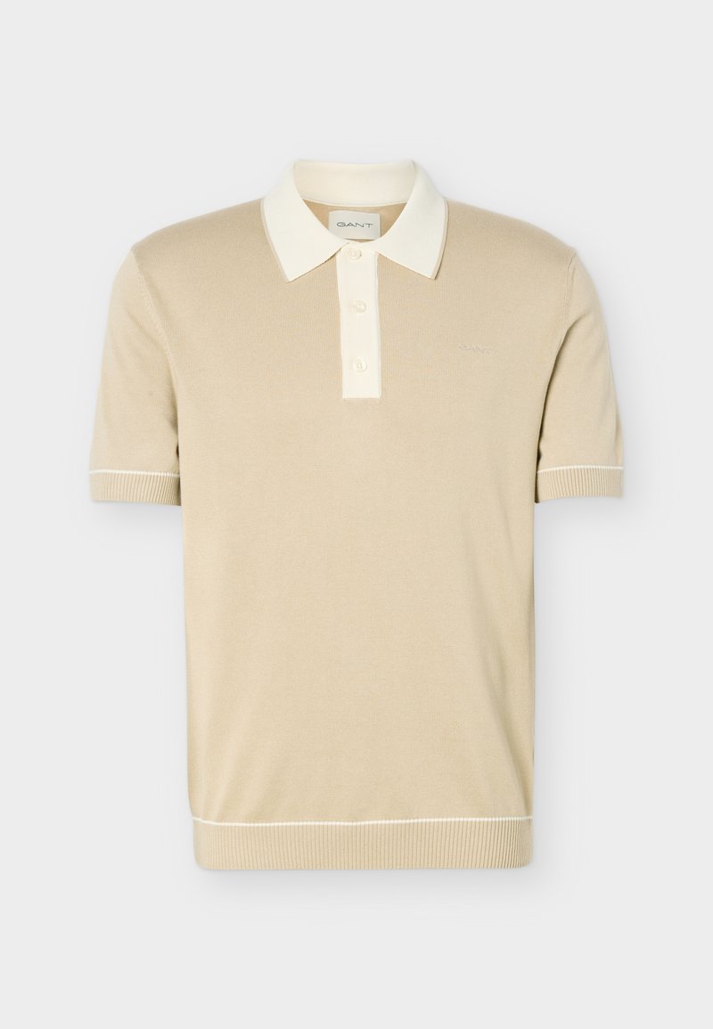 Polo beige à manches courtes avec col blanc, patte de boutonnage à trois boutons, et poignets ainsi que bord inférieur côtelés avec une discrète bordure blanche.