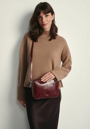 Bordowa torba na ramię ze skóry o błyszczącej fakturze, prostokątnym kształcie, zamkiem błyskawicznym i regulowanym paskiem, noszona z beżowym swetrem.