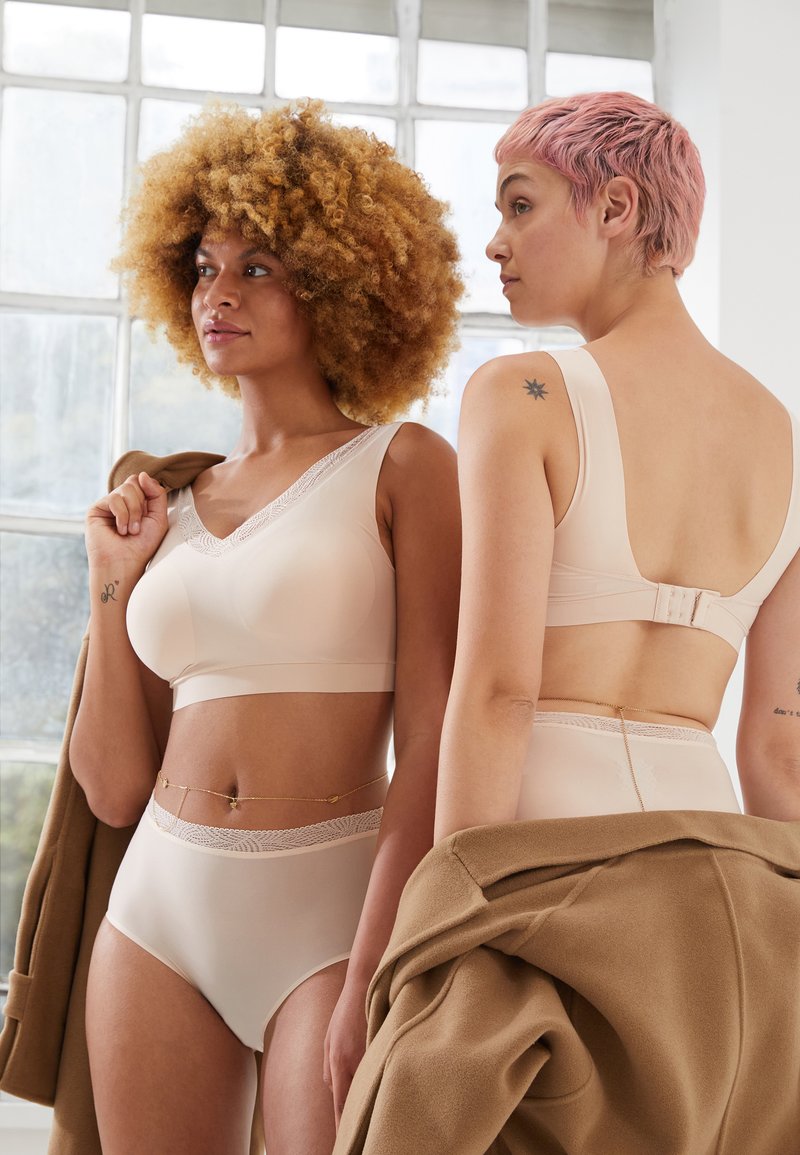 Dos modelos luciendo sujetadores beige sin costuras con encaje y ropa interior de cintura alta, una de ellas lleva un abrigo marrón sobre el hombro.