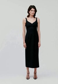 Svart ärmlös klänning med V-ringning, figurnära bodice och rak midi-längd kjol. Slätt tyg, minimal detaljer, parat med remsandaler.