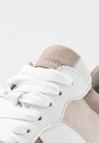 Gros plan sur une basket blanche et beige montrant un cuir texturé, des détails en suède, des lacets blancs et des coutures sur un fond blanc immaculé.