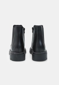 Un paio di stivali neri in pelle alla caviglia con suole in gomma spessa e cerniere posteriori su uno sfondo bianco semplice.