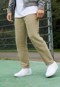 Beige bomullschinos med rak passform, i kombination med vita lädersneakers. I bakgrunden syns en grön yta och ett metallstängsel.