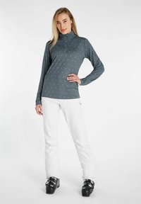 Langsleeve, rits-kraag, grijs topje met kleine, lichtgrijze driehoekige patronen. Gecombineerd met witte broek en zwarte skibotten.