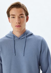 Sudadera azul claro confeccionada en tejido suave, que cuenta con un bolsillo frontal tipo canguro y capucha con cordón. Diseño liso con una textura suave.