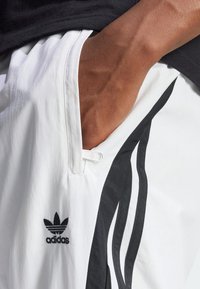 Vita sportbyxor med svarta sidosträck, synlig tygstruktur, med en Adidas-logotyp på vänster höft och en ficka med dragkedjestängning.