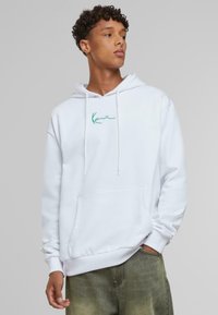 Sudadera blanca de algodón, con un bolsillo frontal, cordones y un logo verde bordado en el pecho. Textura suave.