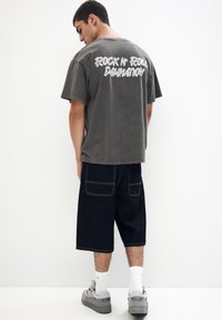 Siva oversize majica s belim napisom "Rock N' Roll Damnation" na hrbtu, v kombinaciji s temnimi denim kratkimi hlačami in sivimi teniskami.