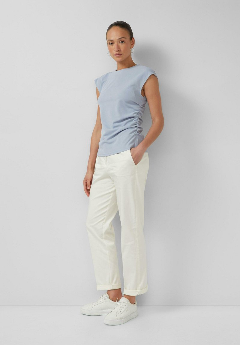 Vrouw staand, gekleed in een lichtblauwe mouwloze top met zijsamendrukking, witte rechte broek, witte sneakers en creolen, tegen een witte muur.