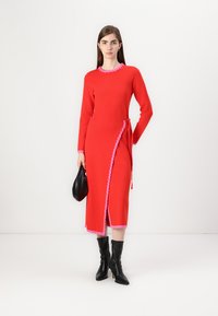 Rotes Strickkleid mit einem pinken Rüschenausschnitt und seitlichem Bindeband. Verfügt über einen knielangen Schlitz und einen kontrastierenden pinken Saum. Kombiniert mit schwarzen Stiefeln und einer Tasche.