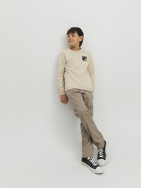 Beige huppari, jossa on musta grafiikka rinnalla, yhdistettynä vaaleanruskeisiin housuihin ja mustiin korkeisiin lenkkareihin, nojaamassa valkoista seinää vasten.