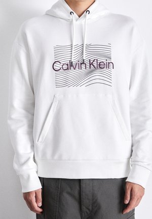 Osoba nosící bílou mikinu s kapucí, přední kapsou, černými vlnitými čarami a nápisem "Calvin Klein" přes hruď.