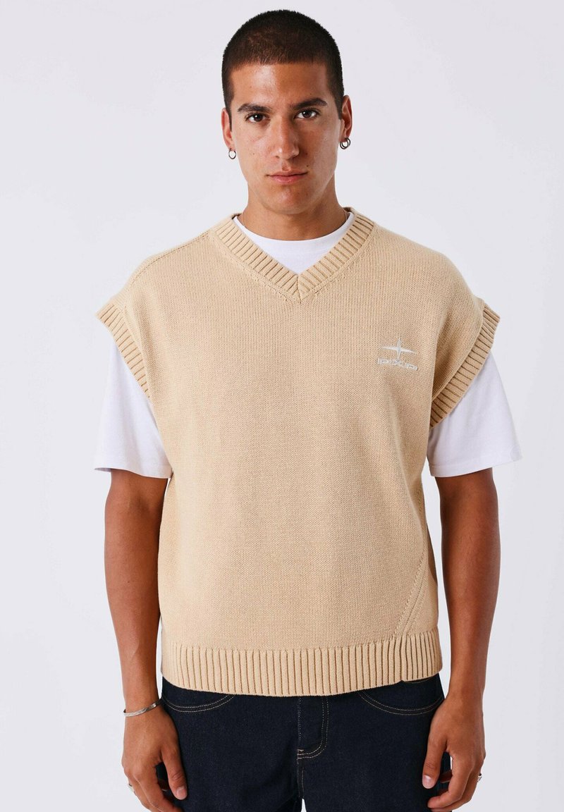 Joven vistiendo un suéter sin mangas de punto beige sobre una camiseta blanca, jeans oscuros y pequeños pendientes de aro, de pie contra un fondo liso.