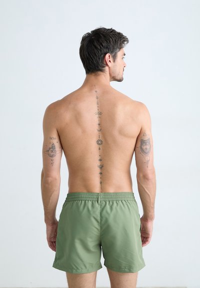 Shorts de bain verts en matériau léger, avec une taille élastique et une texture lisse. Tatouages visibles sur le dos et les bras.