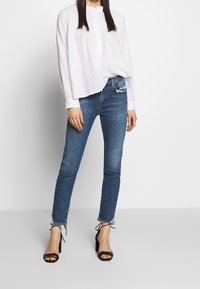 Blusa blanca de manga larga con pliegues, combinada con jeans skinny de color azul desgastado que presentan un dobladillo deshilachado y sandalias de tacón alto negras con tiras.