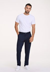 Maglietta bianca a girocollo abbinata a pantaloni eleganti blu navy. I pantaloni hanno un davanti piatto, passanti per la cintura e una chiusura con un solo bottone. Indossati con sneakers bianche.