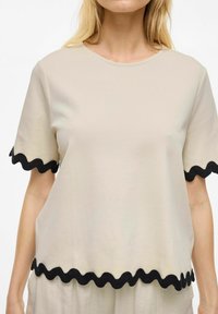 Beige shirt met korte mouwen en een ronde halslijn, afgewerkt met golvende zwarte bies langs de mouwen en onderkant. Zachte stof met een gladde textuur.