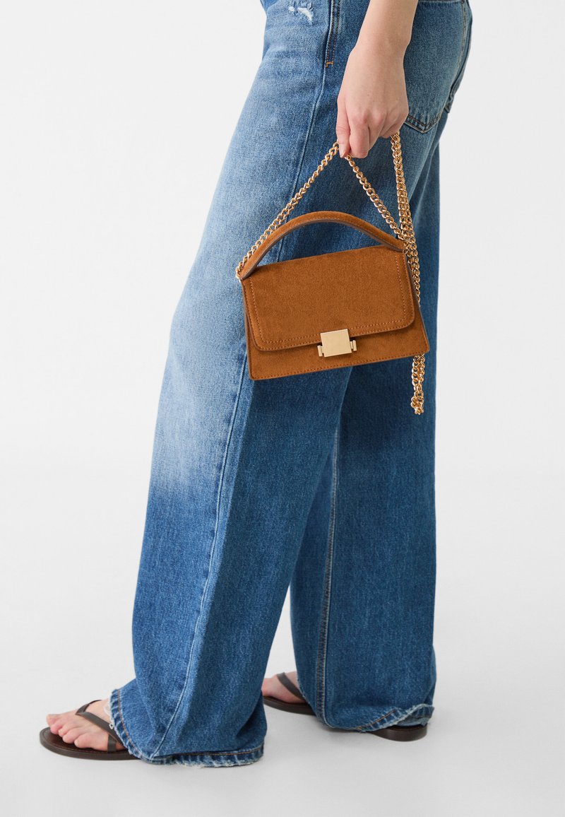 Brown suede handbag with a structured shape, chain strap, and gold hardware. Paired with light blue wide-leg jeans and black sandals.