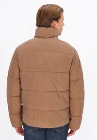 Bruine pufferjack met een hoge kraag en doorgestikte quiltpatroon. Zachte textuur en afgeronde silhoët. Draagt met donkere spijkerbroeken.