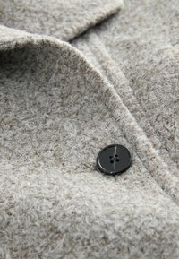 Vaaleanharmaa teksturoitu villakangas, jossa on pehmeä ja karvainen ulkonäkö; reunassa on musta nappi, jossa on neljä reikää.