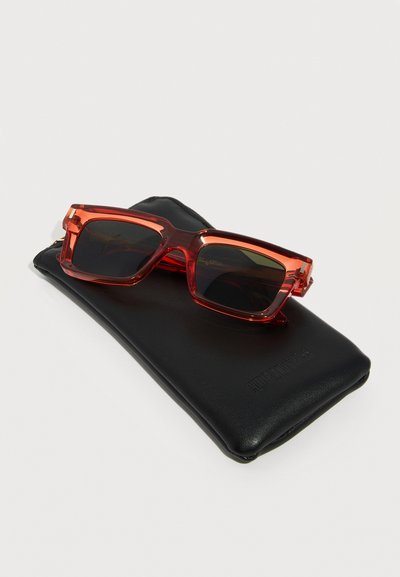 Ορθογώνια πορτοκαλί ημιδιαφανή γυαλιά ηλίου με σκουρόχρωμους φακούς, τοποθετημένα πάνω σε μαύρη δερμάτινη θήκη, σε απλό φόντο.